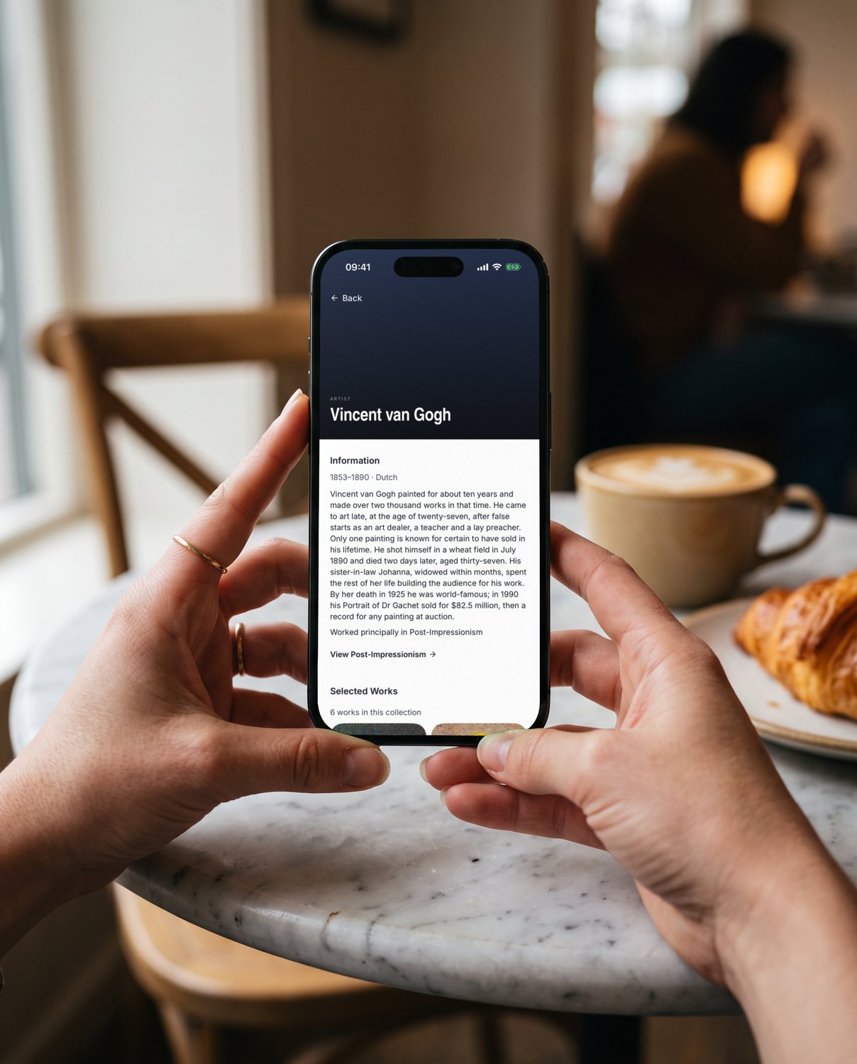 Two hands holding an iPhone in a café, showing Vincent van Gogh's artist page in YEUX.